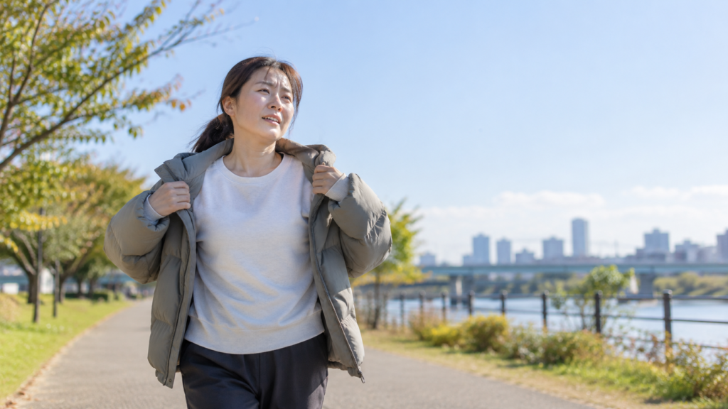 晴れた川沿いを歩きながら、厚手のアウターの前を開けて暑さに合わせて着方を調整する女性